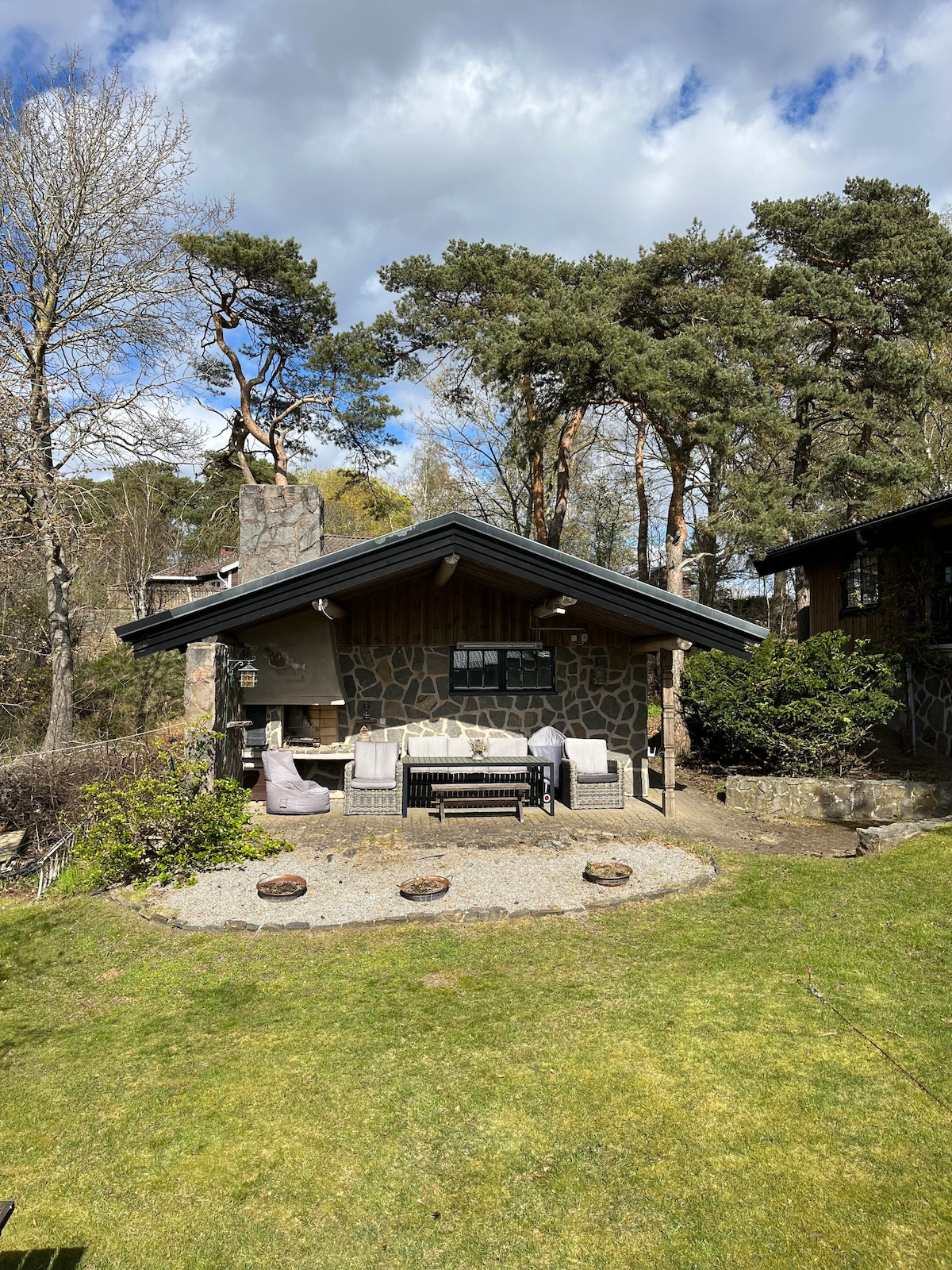 A charming wooden house is set against a backdrop of trees, featuring a stone patio area. Comfortable seating is arranged around a small fire pit, while a well-kept lawn frames the outdoor space, enhancing the natural surroundings.