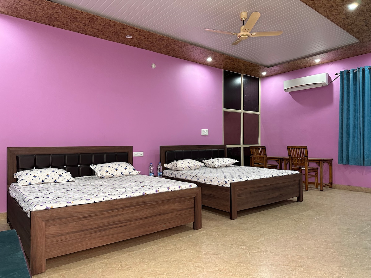 A bright guest room features two spacious beds with patterned linens, positioned against vibrant pink walls. A small dining area with wooden chairs is visible in the background. Natural light enters through a window covered with dark curtains, and a ceiling fan is mounted above.