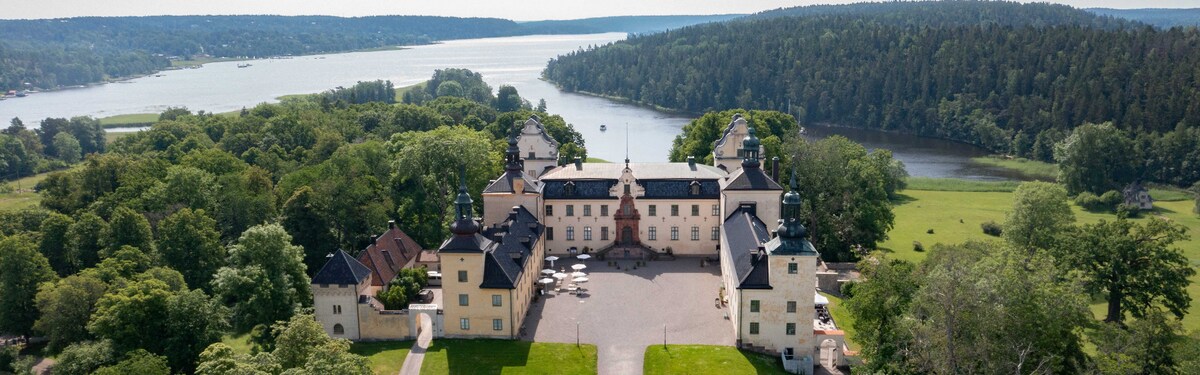 An aerial view of a large historic building surrounded by lush greenery and water. The structure features multiple turrets and a spacious courtyard with tables. The scenic landscape includes a river and forested hills in the background, creating a serene atmosphere.