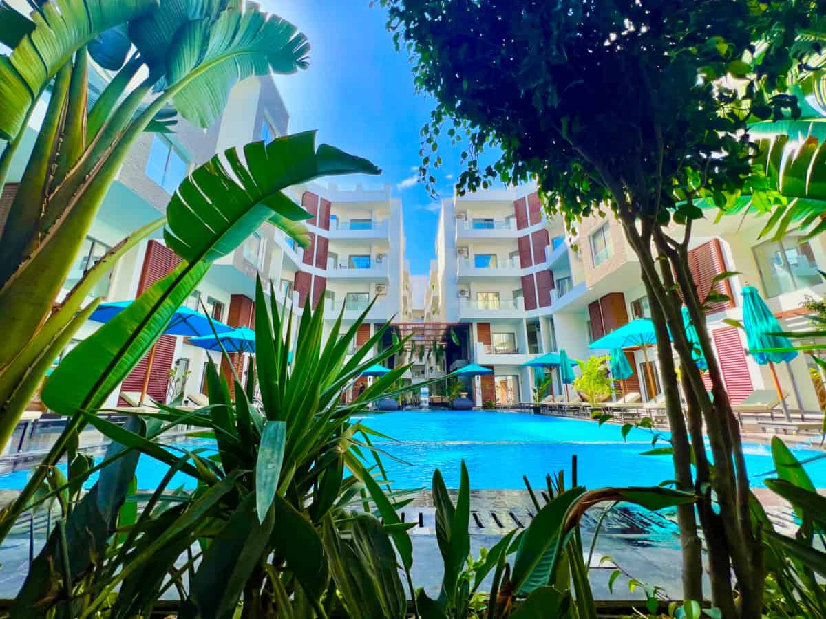 A view is captured from a garden area, showcasing a pool surrounded by modern buildings. Lush green plants frame the foreground, while bright umbrellas provide shade around the poolside. The clear blue sky complements the inviting atmosphere of the recreation area.