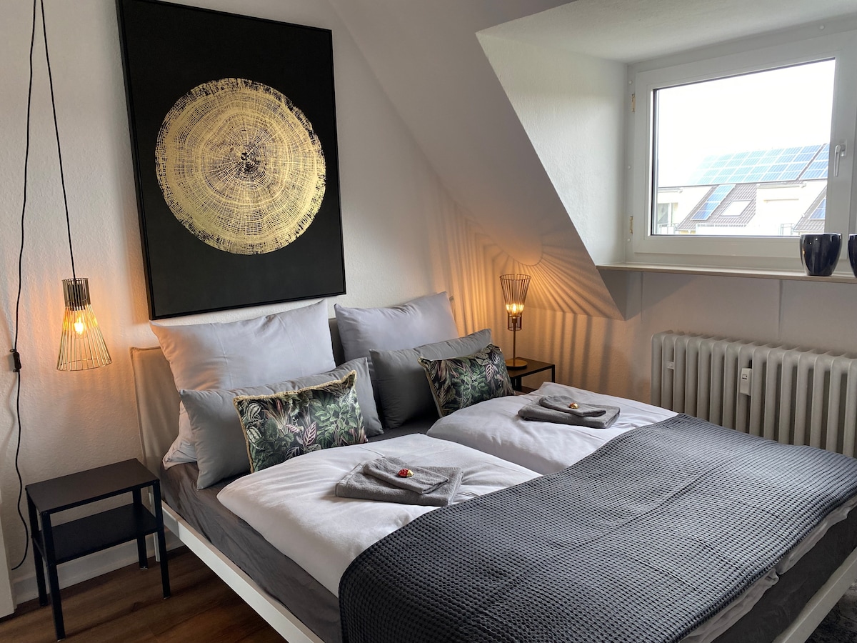 A well-appointed sleeping area features a spacious bed with gray blankets and decorative pillows. Soft lighting is provided by two bedside lamps, with a modern artwork displayed above the bed. A window allows natural light to illuminate the room.