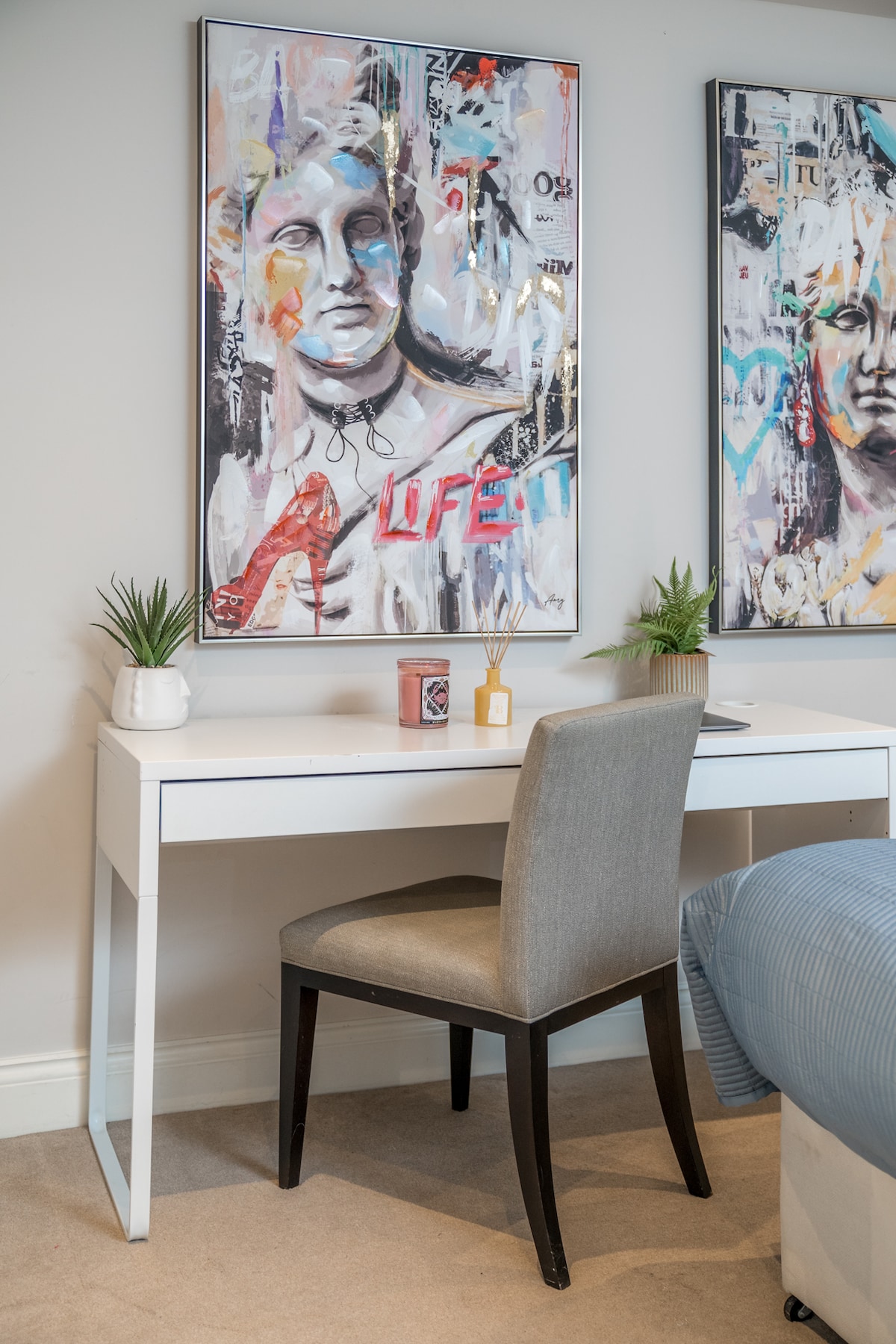 An inviting workspace is showcased with a sleek white desk complemented by a comfortable grey chair. Two small potted plants sit on the desk, while vibrant artwork adorns the wall above, adding a contemporary touch to the room's decor.