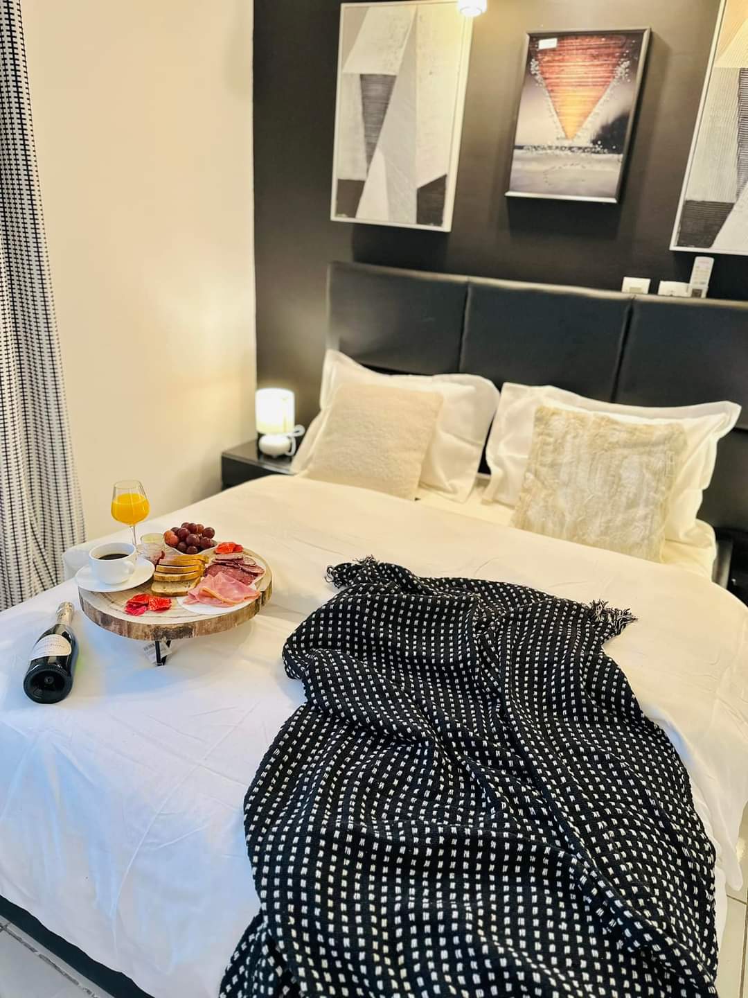 A serene sleeping area features a spacious bed with plush pillows and a textured throw draped across it. A wooden tray holds a selection of fruits, snacks, and a glass of orange juice. Abstract wall art is displayed above the bed, enhancing the modern decor.