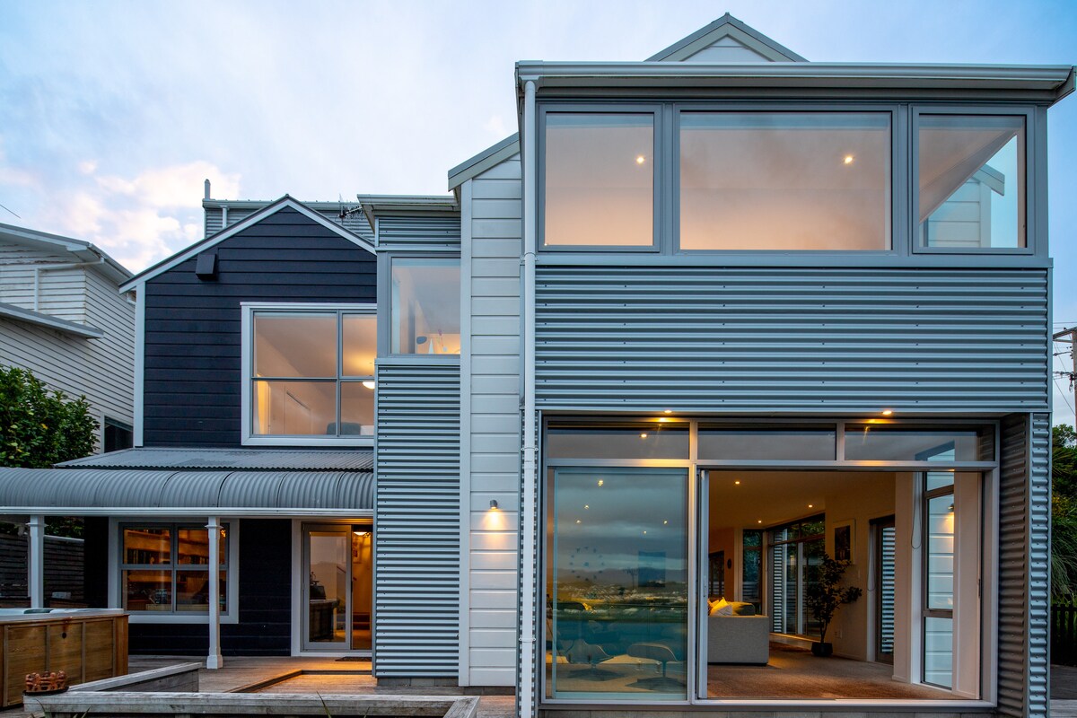 The two-story house features a modern design with a combination of dark and light siding. Expansive glass windows and doors provide ample natural light, while a spacious outdoor area is visible, inviting connection with the surrounding views.