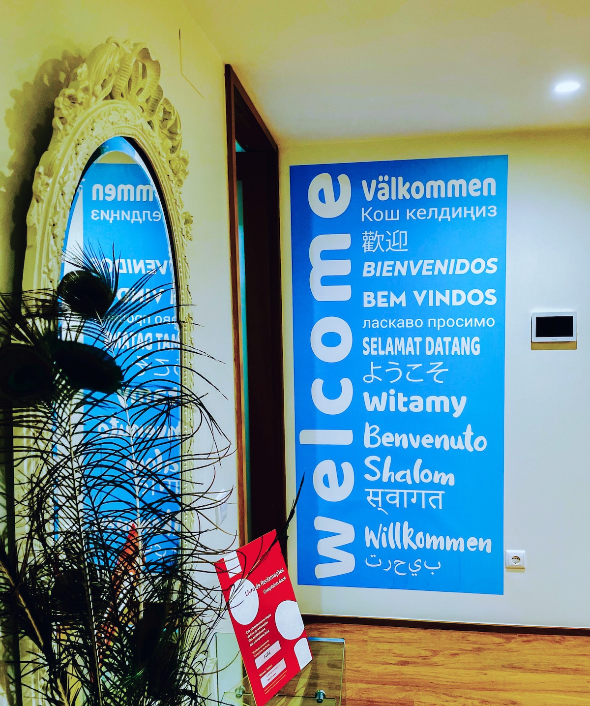 A welcoming entrance area is highlighted by a prominent wall featuring the word 'welcome' in various languages. A large decorative mirror reflects the space, complementing the minimalist design of the foyer.