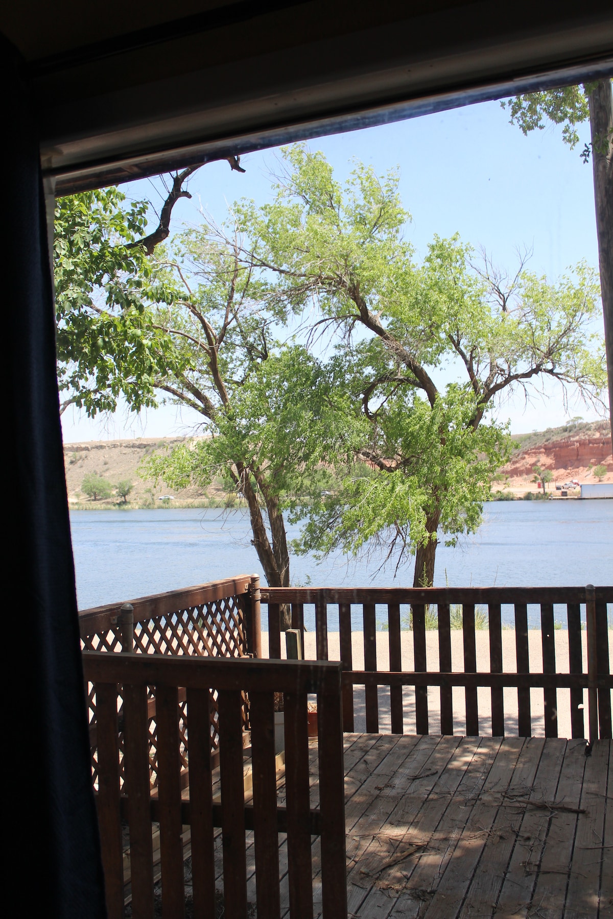 A view from the window showcases a wooden deck overlooking a serene lake. Lush green trees frame the scene, creating a natural border. The calm water reflects the clear blue sky, offering a peaceful and inviting outdoor environment.