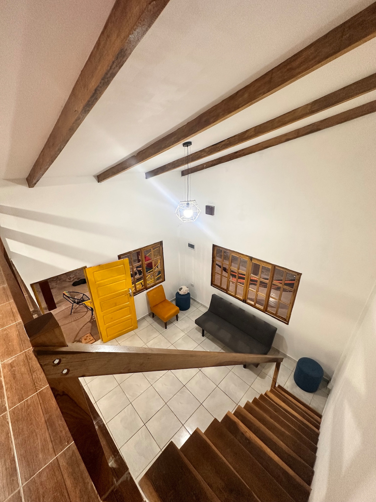 An overhead view of a spacious living area is presented, featuring wooden beams across a bright ceiling. A dark sofa is positioned next to an orange cabinet, with a blue accent chair nearby. Large windows allow natural light to filter into the room.