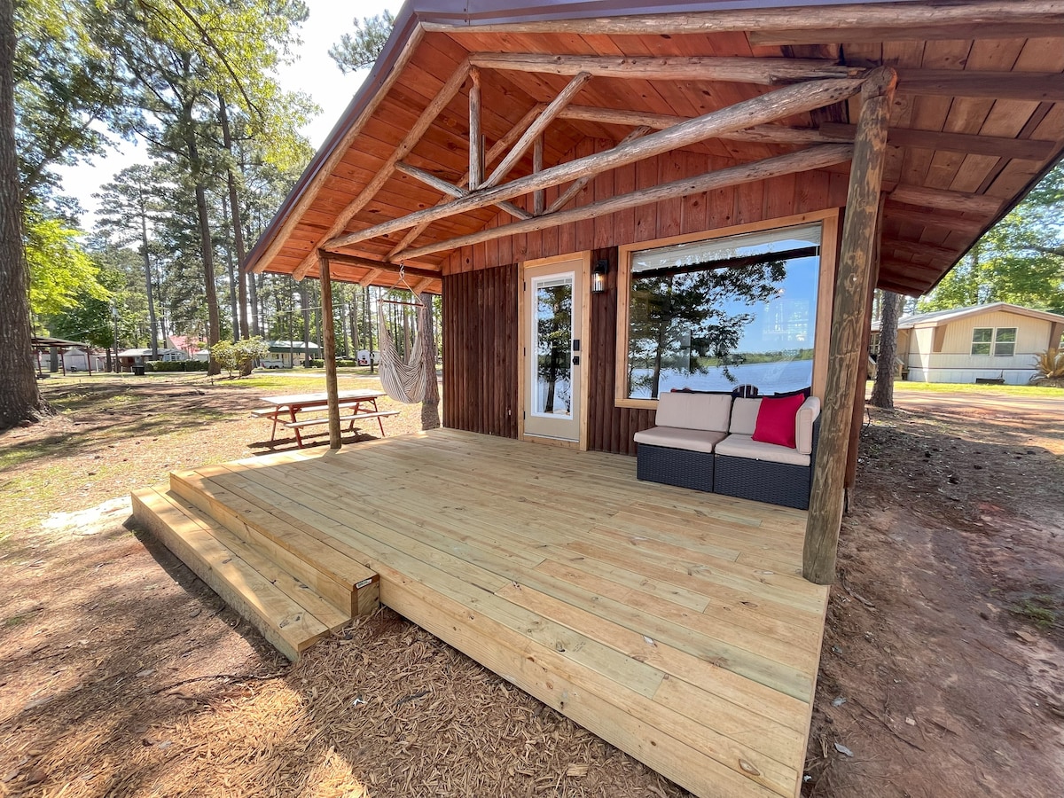 A cedar cabin exterior is presented, featuring a covered porch with a comfortable seating area and a picnic table nearby. Large windows provide views of the surrounding nature, while wooden steps lead up to the entryway.