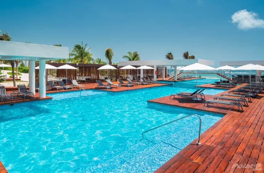 A large outdoor pool is surrounded by wooden decking, featuring multiple lounge chairs with umbrellas for shade. Palm trees and clear blue skies are visible in the background, enhancing the serene atmosphere of the space.