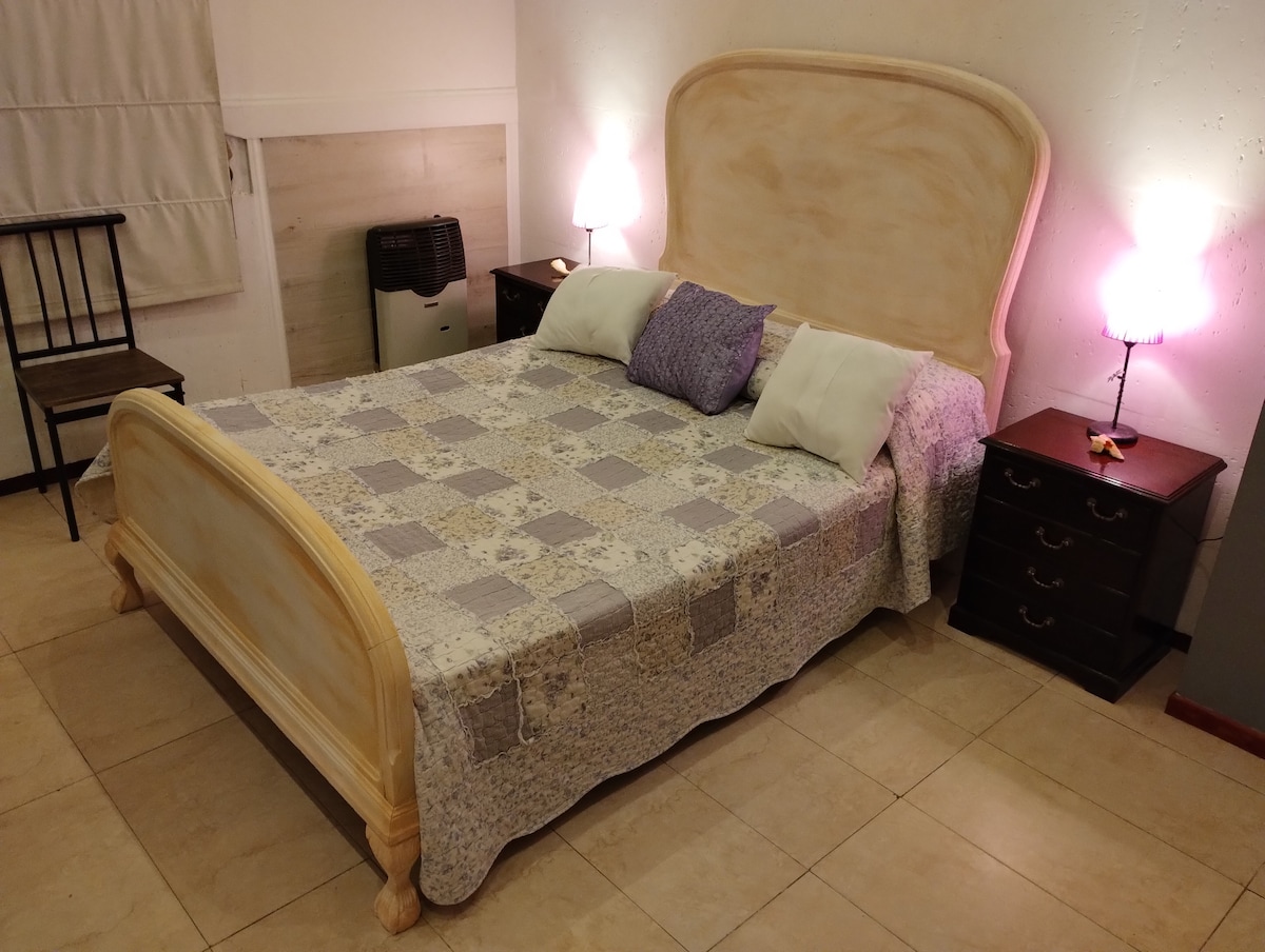 A spacious bed is positioned between two nightstands, featuring bedside lamps that provide soft lighting. The bed is dressed with a multi-colored quilt and complemented by decorative pillows. A simple chair and a small table are visible in the background.