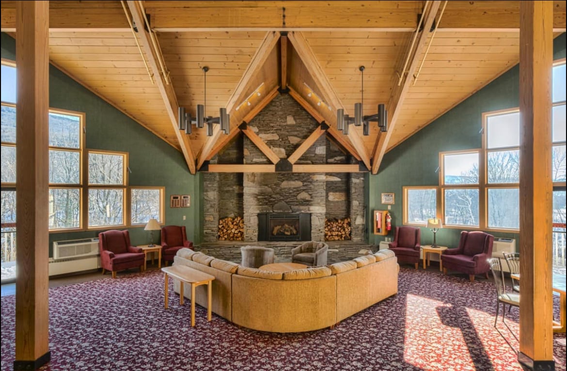 A spacious lounge area is captured, featuring a large circular sofa and several comfortable armchairs. A stone fireplace serves as a central focal point, framed by vaulted wooden ceilings and large windows that provide natural light and views of the surrounding landscape.