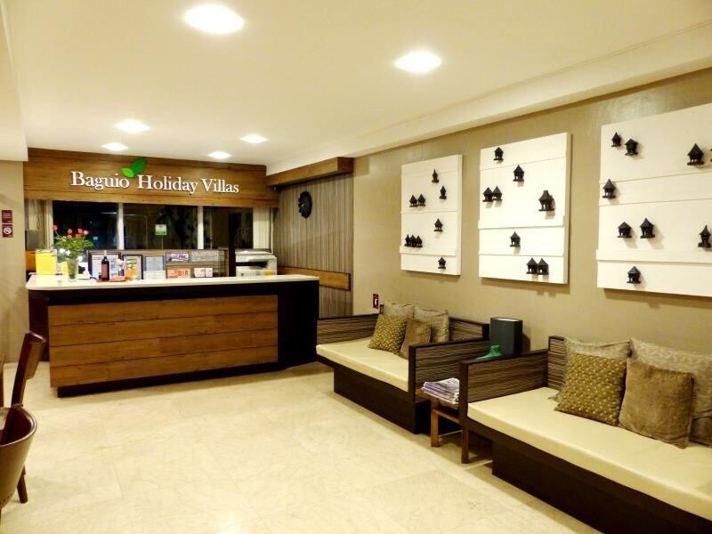 The reception area features a large wooden desk with a modern design. Comfortable seating is provided along the walls, adorned with decorative panels showcasing small house silhouettes. Soft lighting enhances the welcoming atmosphere, while large windows allow natural light to fill the space.
