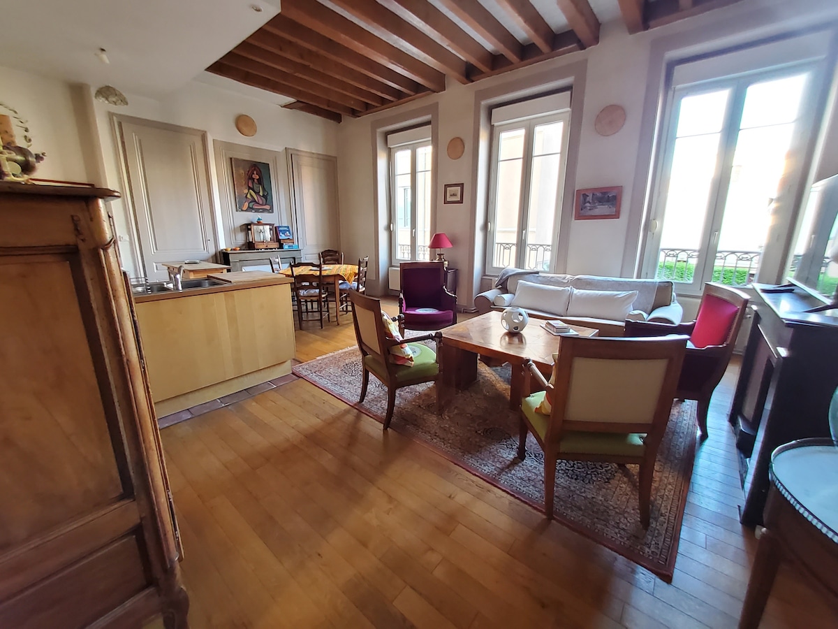 A spacious living area showcases wooden beam ceilings and natural light from large windows. The room contains a dining table with chairs, a cozy sofa, and a couple of armchairs, creating a welcoming space for relaxation and conversation. A decorative rug enhances the warmth of the hardwood floor.