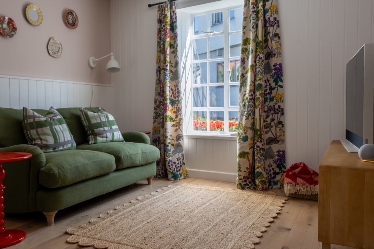 A cozy lounge area features a green sofa adorned with checked cushions. Floral-patterned curtains frame a window that allows natural light to illuminate the room. A decorative rug rests on the wooden floor, and a modern television is positioned on a wooden console.