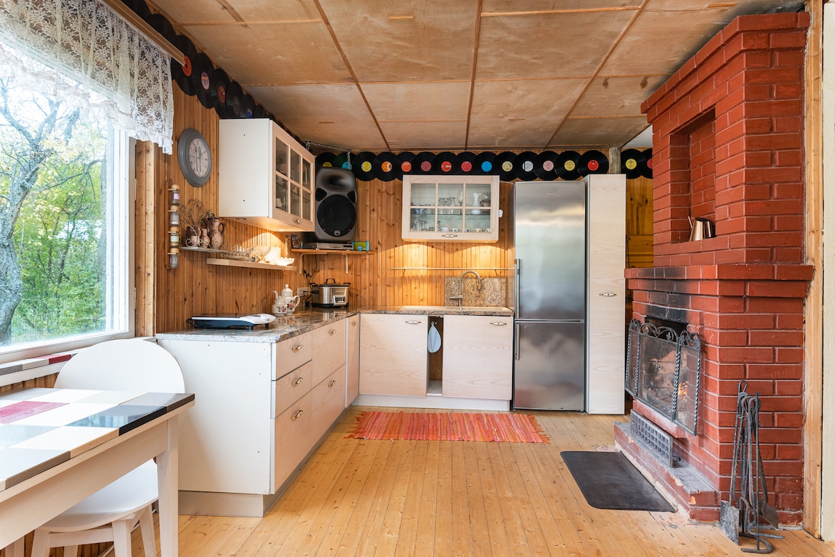The kitchen is equipped with modern appliances, including a refrigerator and oven. Natural light enters through a large window, illuminating the wooden cabinetry and countertops. A small dining area with a simple table and chairs is situated nearby, with a brick fireplace adding warmth to the space.