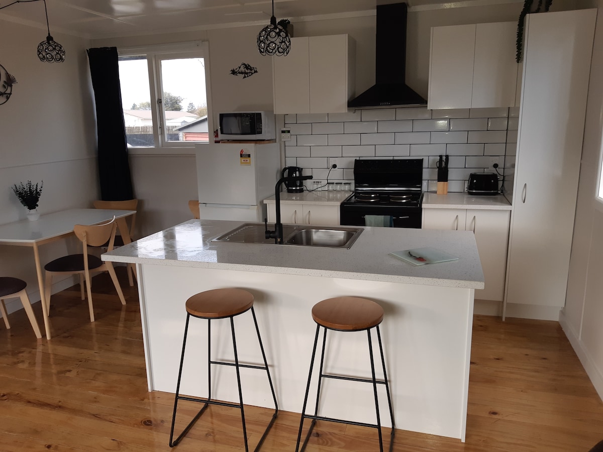 A modern kitchen features a central island with two bar stools. Stainless steel appliances are set against white cabinetry and a tiled backsplash. A dining area with light wood chairs is visible, along with large windows allowing natural light to brighten the space.