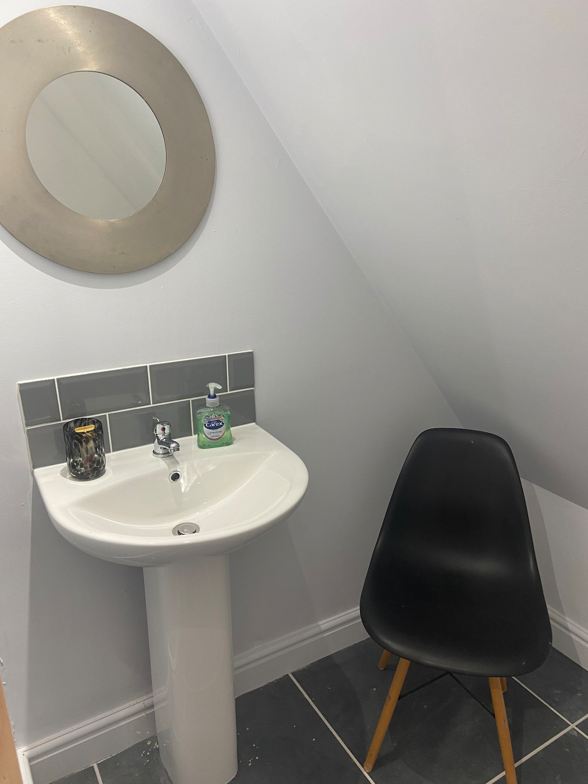 A compact bathroom features a modern pedestal sink with a round mirror above. Gray tiles adorn the wall behind the sink, while a sleek black chair occupies the small space. The floor is tiled in a darker grey, providing a cohesive look.