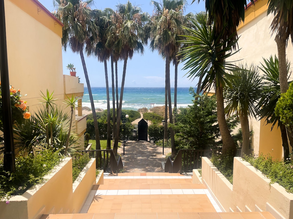 A path lined with tropical palm trees leads towards the beach, with the ocean visible in the distance. Lush greenery surrounds the area, providing a serene backdrop. The entrance to a gated access point is positioned at the end of the pathway, inviting guests to explore the beachfront.