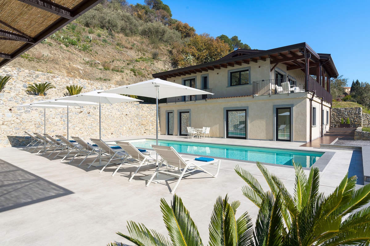 A spacious terrace features a saltwater pool surrounded by lounge chairs and large umbrellas, with the hillside home in the background. The exterior combines pale stone walls with dark beams, complemented by greenery and mountain views.
