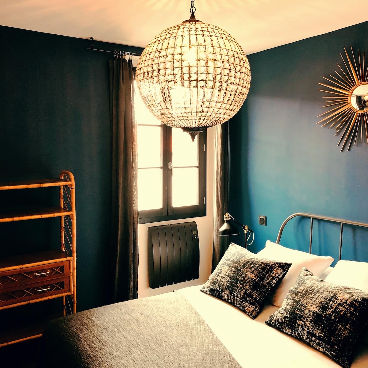A cozy bedroom is highlighted by a large, decorative chandelier, casting a warm glow. A spacious bed with textured pillows faces a window adorned with dark curtains. A wooden shelving unit and a wall mirror complete the inviting space, emphasizing the rich blue walls.