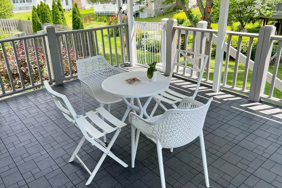 A covered outdoor patio area features a round table surrounded by four white wicker chairs. Lush greenery and colorful flower beds are visible in the background, enhancing the serene outdoor setting. The textured flooring provides a modern touch to the space.