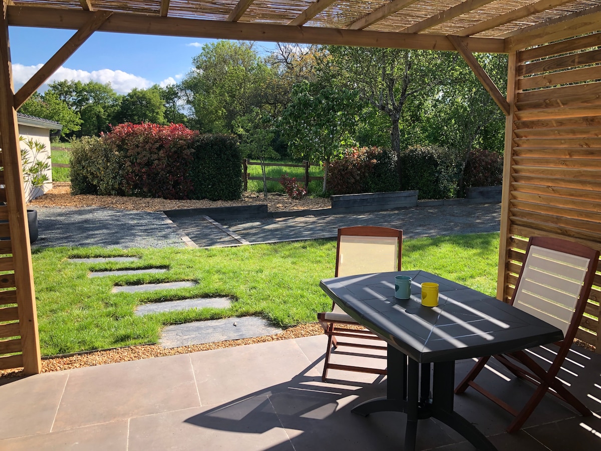 A terrace is situated under a wooden pergola, furnished with a table and chairs. Two colored mugs rest on the table. The view includes a well-maintained garden with grass and shrubs in the background, framed by a pathway made of stones.
