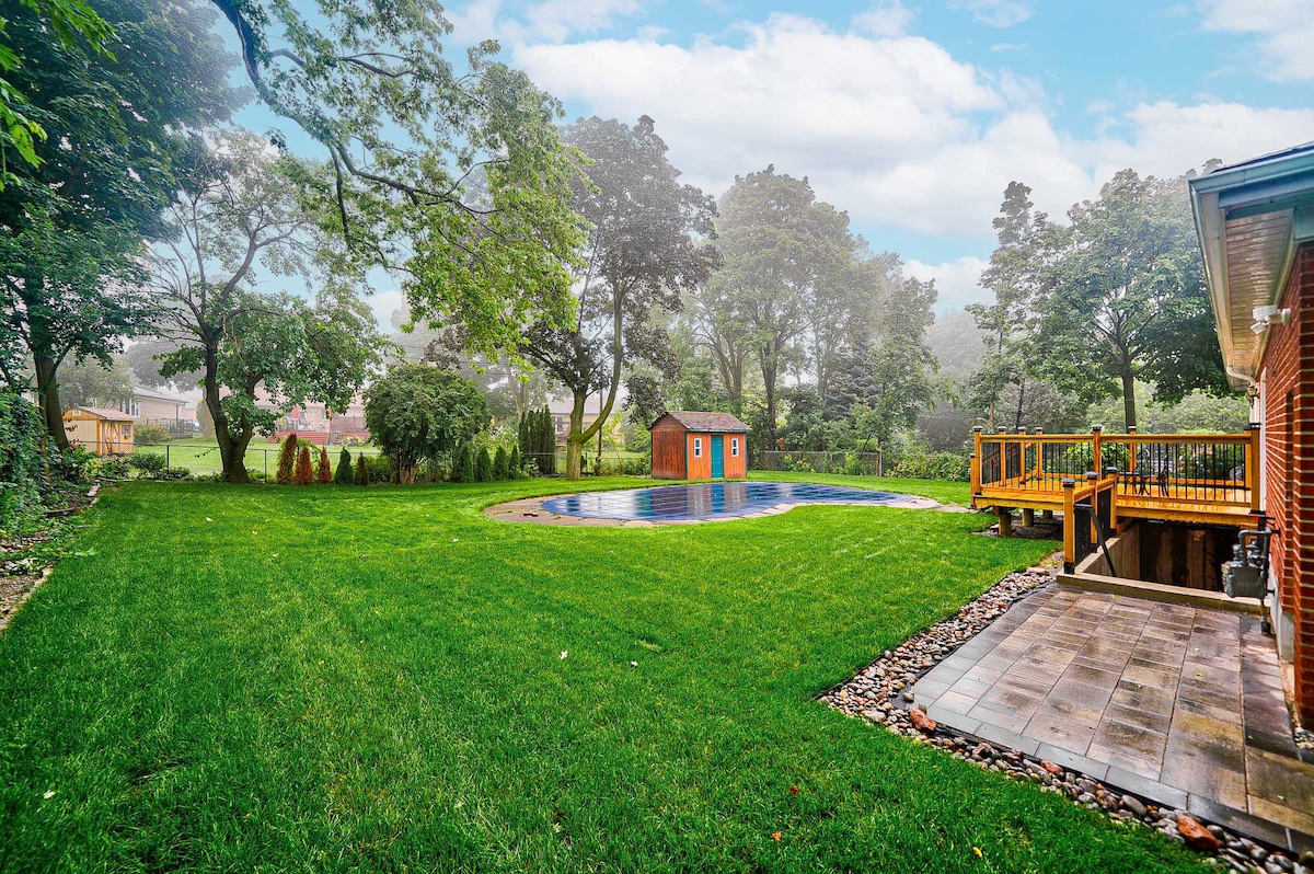 An expansive yard is showcased with a well-maintained lawn surrounding a pool. Two storage sheds are seen in the distance, and a wooden deck is positioned to the right, bordered by a paved area. Towering trees provide a natural backdrop.