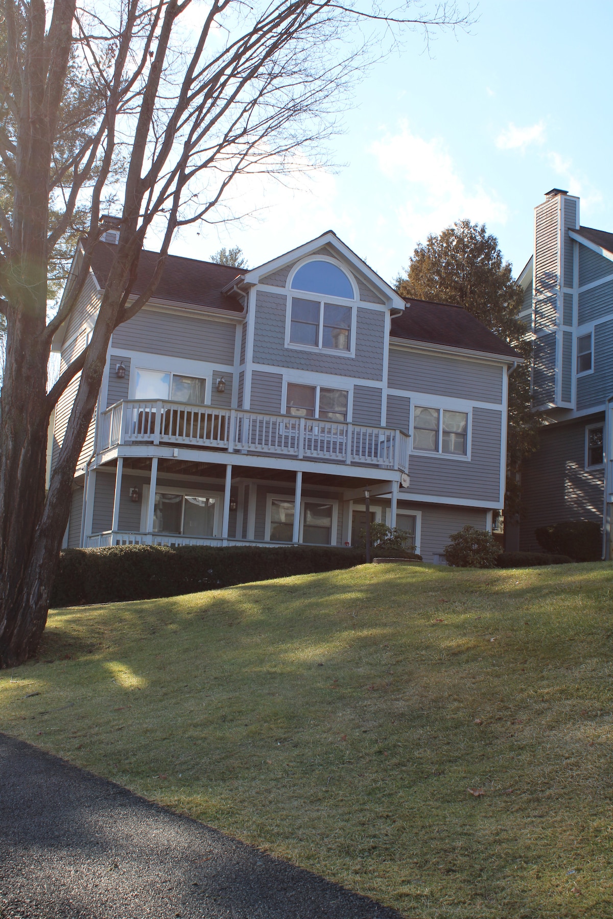 A multi-level property is presented, featuring a light grey exterior and prominent architectural details such as a large arched window. A covered front porch with white railings is visible, surrounded by a neatly maintained lawn and trees, while adjacent homes can be seen in the background.
