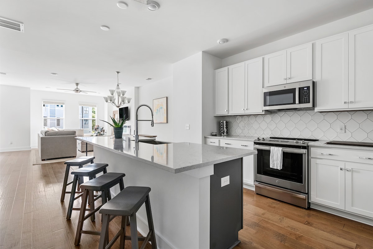 A modern kitchen features sleek white cabinetry and a spacious island with seating for four. Stainless steel appliances, including a microwave and oven, complement the grey countertop. Natural light fills the area, highlighting the open layout that seamlessly connects to the living space.