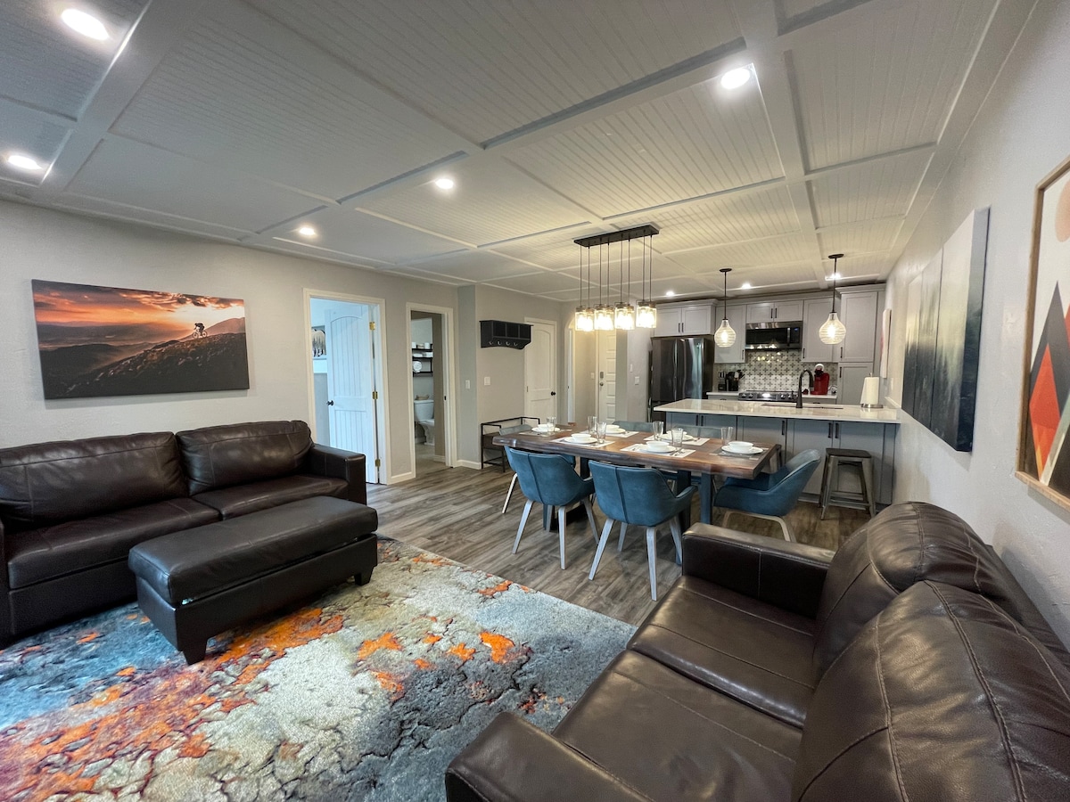 A cozy living space features a dark leather sectional sofa and a vibrant area rug with warm tones. A dining table for six is visible, accompanied by stylish blue chairs. An open kitchen area is seen in the background, with modern lighting and sleek finishes.