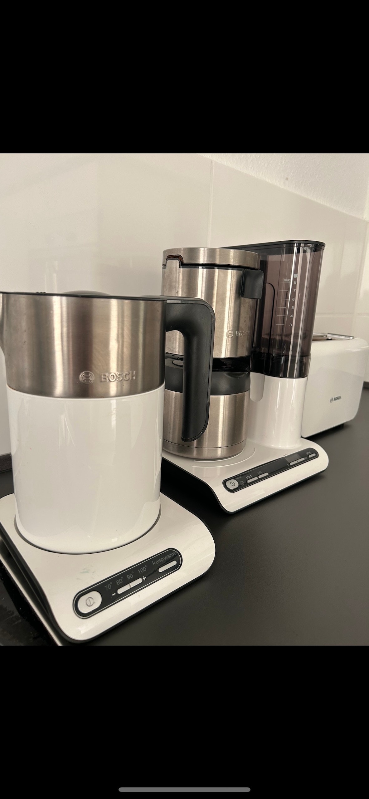 Three modern kitchen appliances are placed on a sleek black countertop. A stainless steel kettle, a programmable coffee maker with a glass carafe, and a white toaster are visible, all designed with a clean and contemporary aesthetic. The surfaces appear organized and inviting for meal preparation.