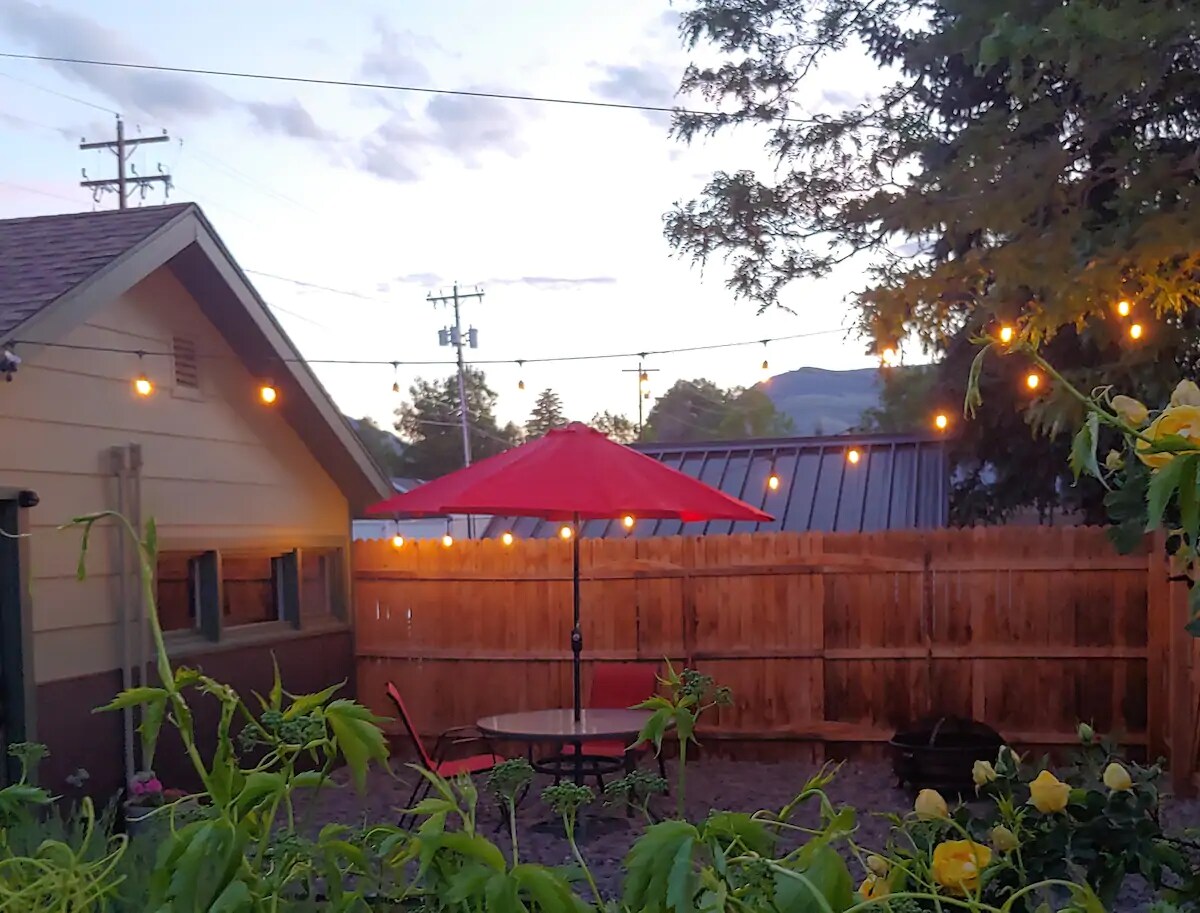 The small private yard features a round dining table with four chairs, shaded by a red umbrella. Warm string lights illuminate the space at dusk, surrounded by a wooden fence. Colorful plants add greenery in the foreground, enhancing the outdoor relaxing area.