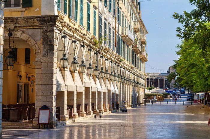 An attractive street in the old town of Corfu is lined with classic architecture, featuring arched walkways and stone columns. Hanging lanterns add charm, while the bright sunlight reflects off the polished pavement. Outdoor seating arrangements are visible, inviting visitors to enjoy the vibrant atmosphere.