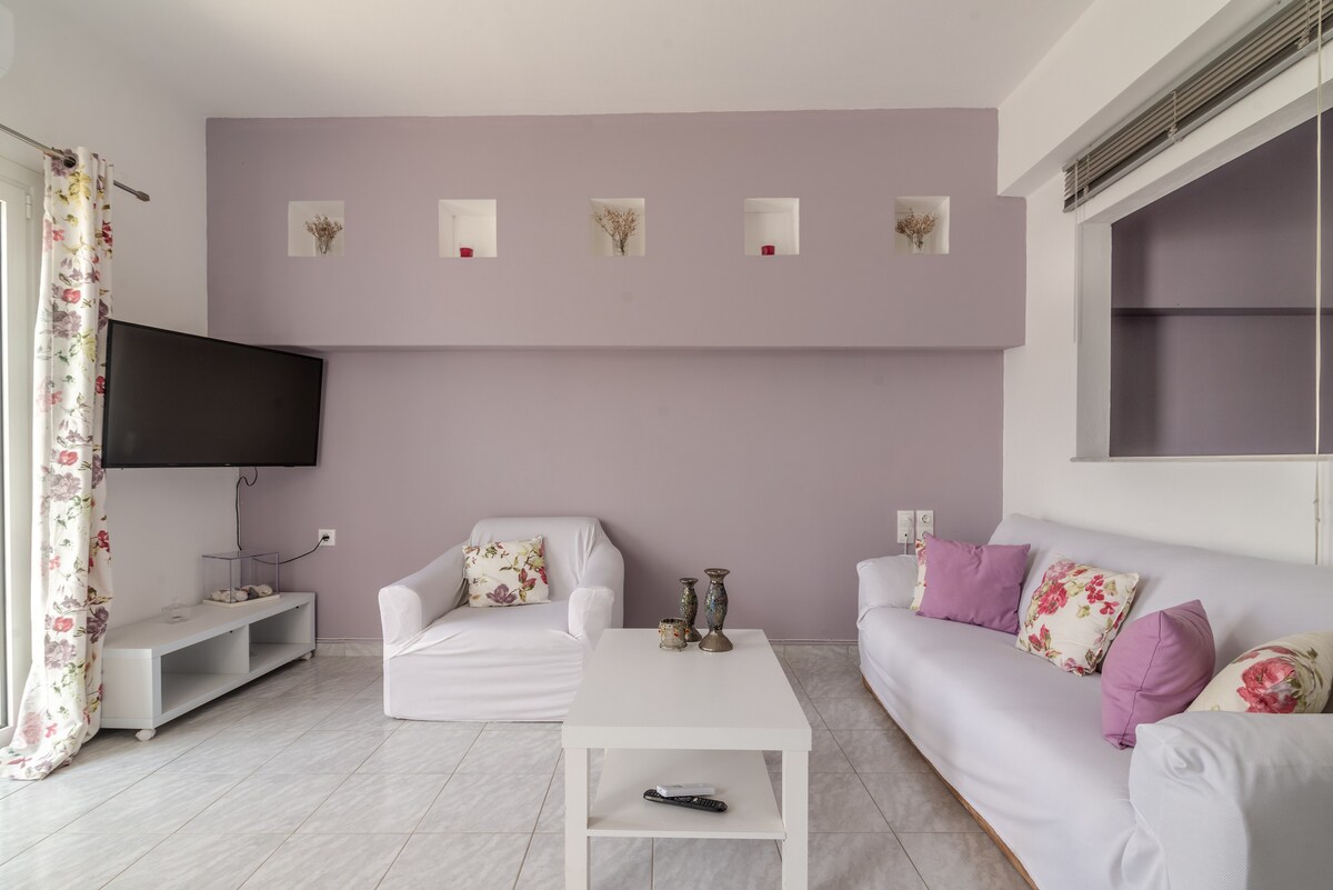 The living room is adorned with soft purple walls and features a comfortable white sofa and armchair, both accented with floral cushions. A white coffee table sits at the center, and a wall-mounted television is positioned above a low entertainment unit.