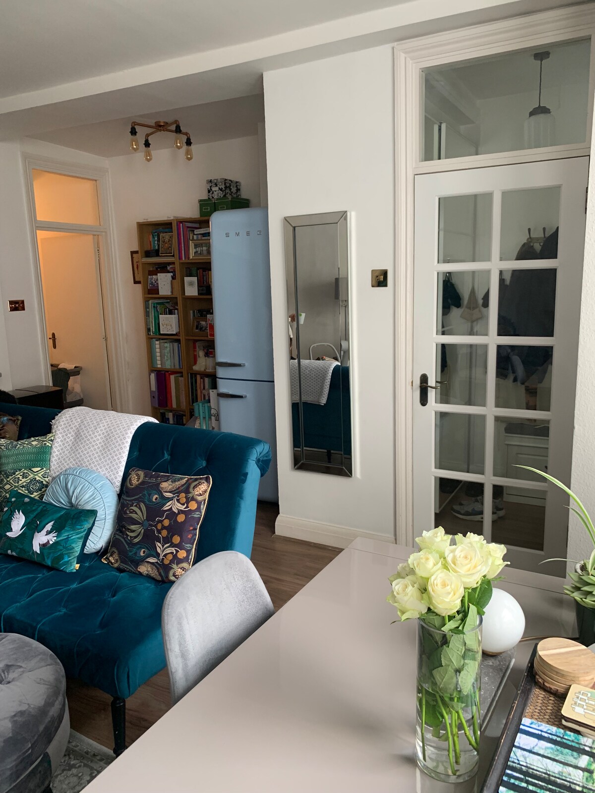 A vibrant living space is presented with plush furniture in shades of blue and gray. A mirrored wall reflects light, creating additional depth. Nearby, a cozy shelving unit displays books and decorative items. Fresh flowers are arranged in a vase on a table, adding a touch of nature.