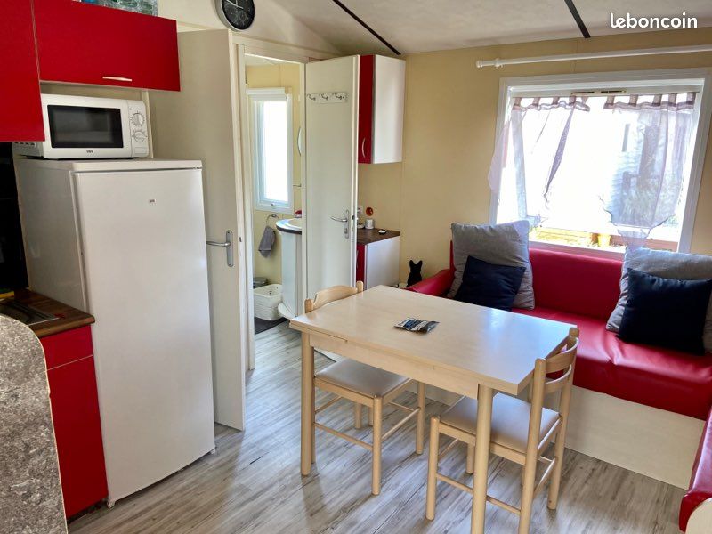 A compact kitchen area features red cabinetry and modern appliances, including a refrigerator and microwave. A light wood table is surrounded by four chairs, with a cozy seating nook visible in the background. Natural light enters through the window, enhancing the space’s comfort.
