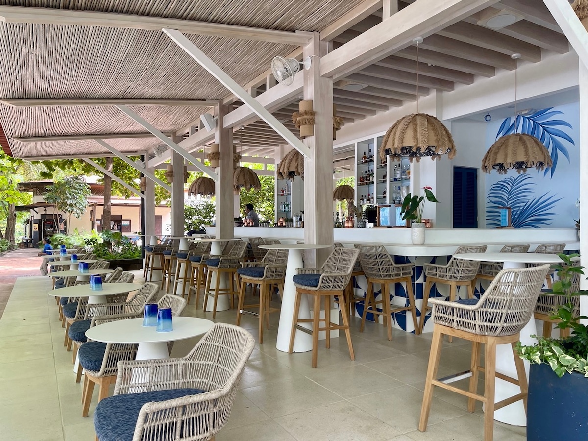 An open-air bar area features a white counter with high chairs surrounded by tropical plants. Unique light fixtures made of natural materials hang overhead, and a blue accent wall adorned with palm leaf designs adds a refreshing touch. Cozy tables with glassware invite relaxation.