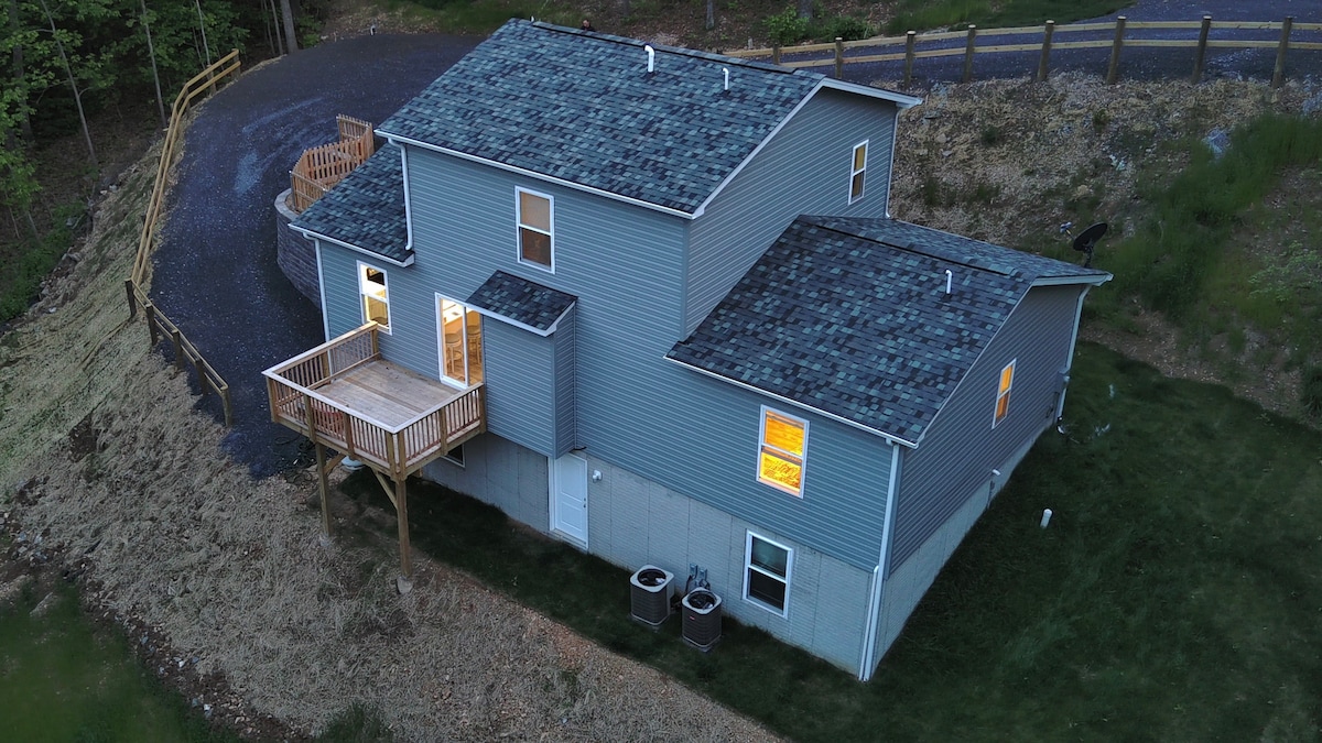 An aerial view captures the three-level modern home surrounded by natural terrain. The back deck is visible, extending from the main structure, with warm light emanating from the windows. Two air conditioning units can be seen near the foundation.