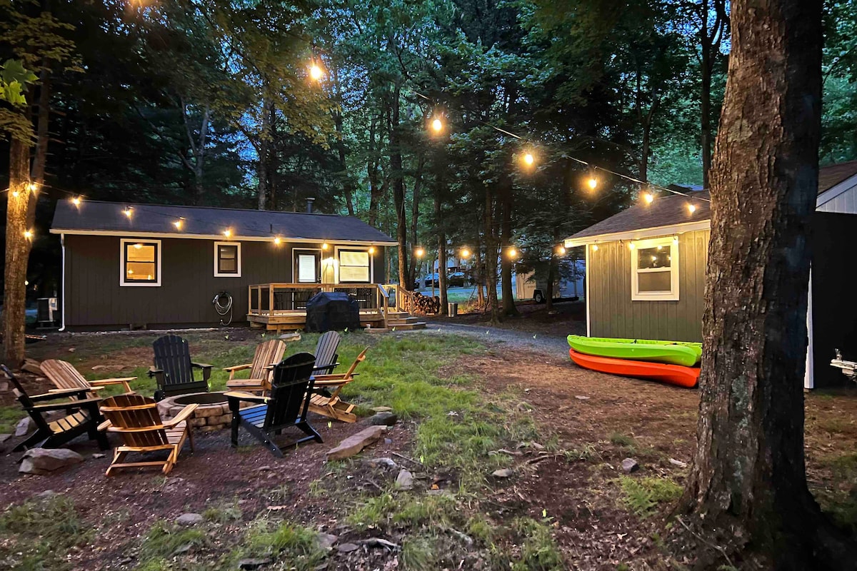 An outdoor area is illuminated by string lights, creating a warm setting. Several wooden Adirondack chairs are arranged around a fire pit. A brightly colored kayak rests on the grass, next to a cozy cabin in a wooded environment.