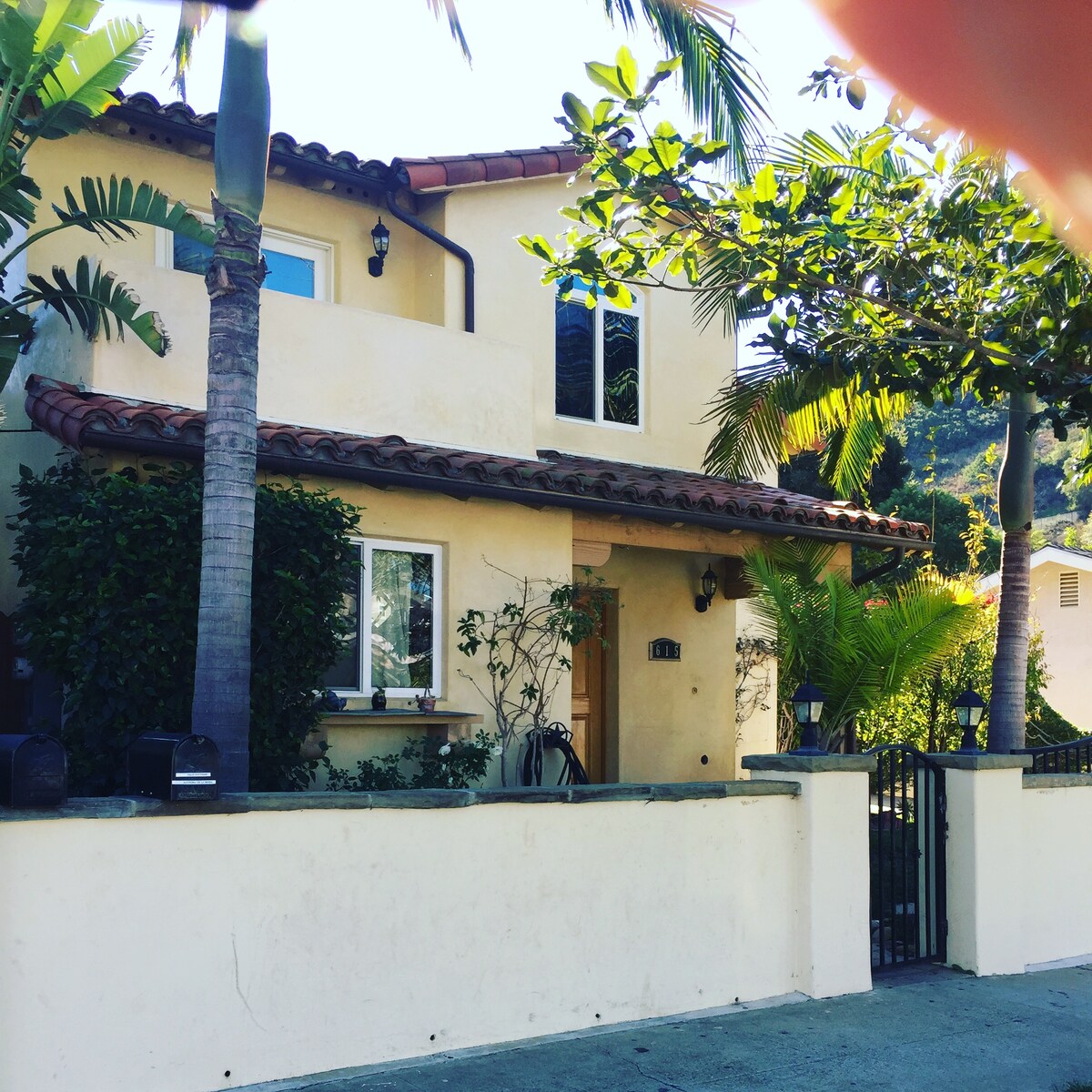 The exterior of a Spanish-style townhouse is shown, featuring warm-toned stucco walls and a terracotta tiled roof. Lush greenery, including palm trees and shrubs, surrounds the entrance, while large windows allow natural light to fill the interior.