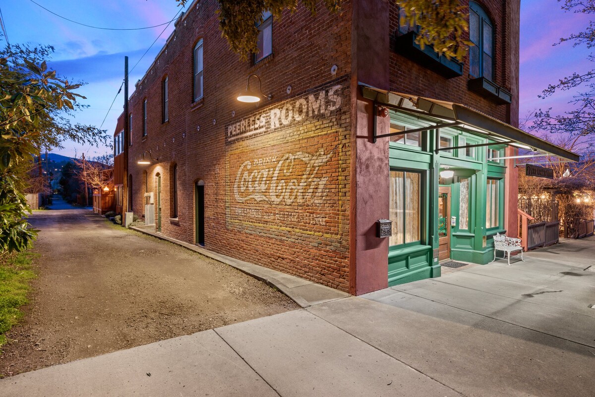 A charming entrance to the historic Peerless Hotel is highlighted by a vintage Coca-Cola mural on the brick exterior. Green doors and large windows complement the rustic brick wall, while pathway lighting and surrounding greenery enhance the inviting atmosphere.