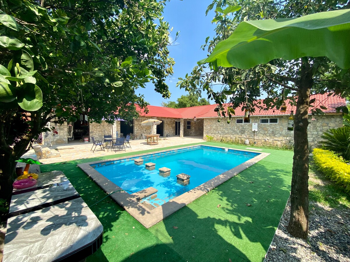 A clear blue pool is surrounded by landscaped greenery and stone pathways. Lounge chairs are positioned near the water, with a shaded seating area under an umbrella. The rustic stone structure with a red roof is visible in the background, complemented by lush foliage.