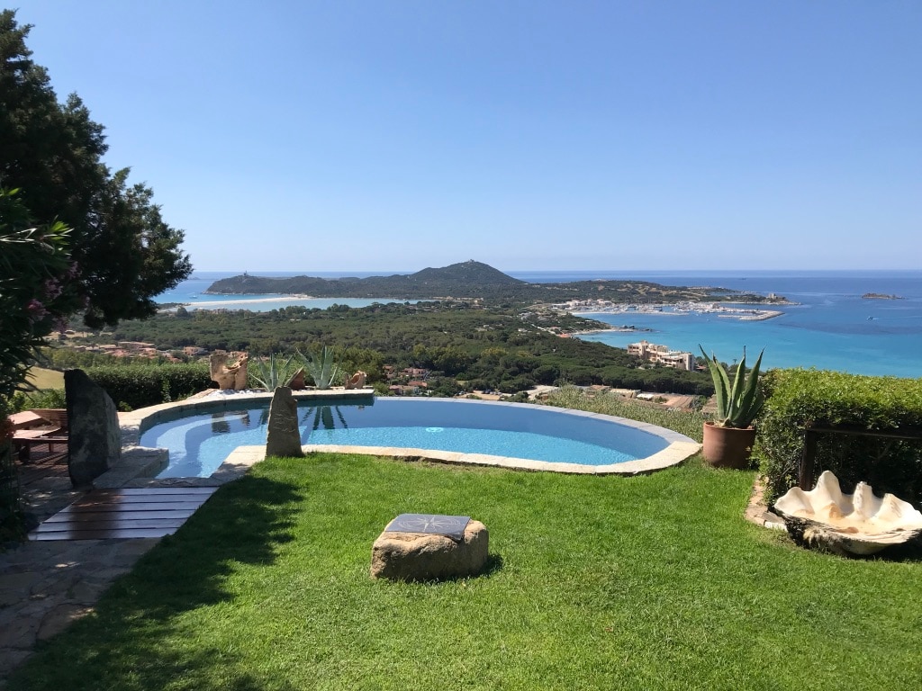 A private pool sits within a well-maintained garden, offering panoramic views of the surrounding coastline and lush landscape. The area features stone accents and various plants, enhancing the serene outdoor setting. Clear blue skies complement the inviting water, merging seamlessly with the horizon.
