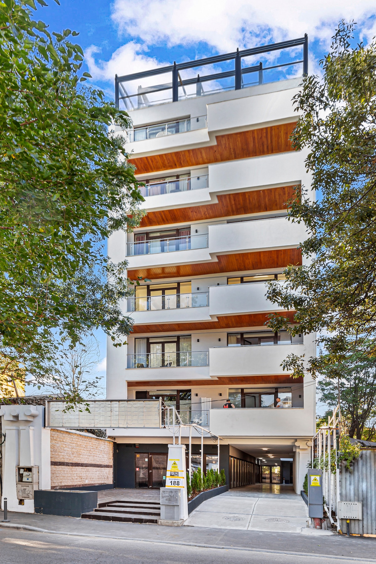 A modern multi-story building is framed by lush green trees, showcasing a sleek facade with wooden accents. Balconies line each floor, allowing natural light to fill the spaces. An entrance is visible at the ground level, leading to a well-maintained pathway.