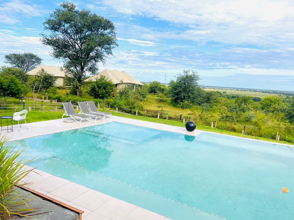 A tranquil swimming pool is visible, surrounded by lush greenery. Two lounge chairs are arranged poolside, offering a space to relax. A distant view of accommodation units is framed against a bright sky, enhancing the serene atmosphere.
