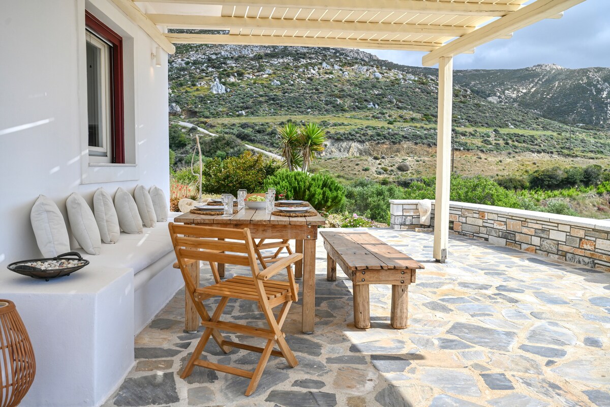 A spacious outdoor patio features a wooden dining table surrounded by folding chairs. A cushioned bench runs along the wall, complemented by stone flooring. The area is framed by a pergola, providing shade while overlooking lush green hills.