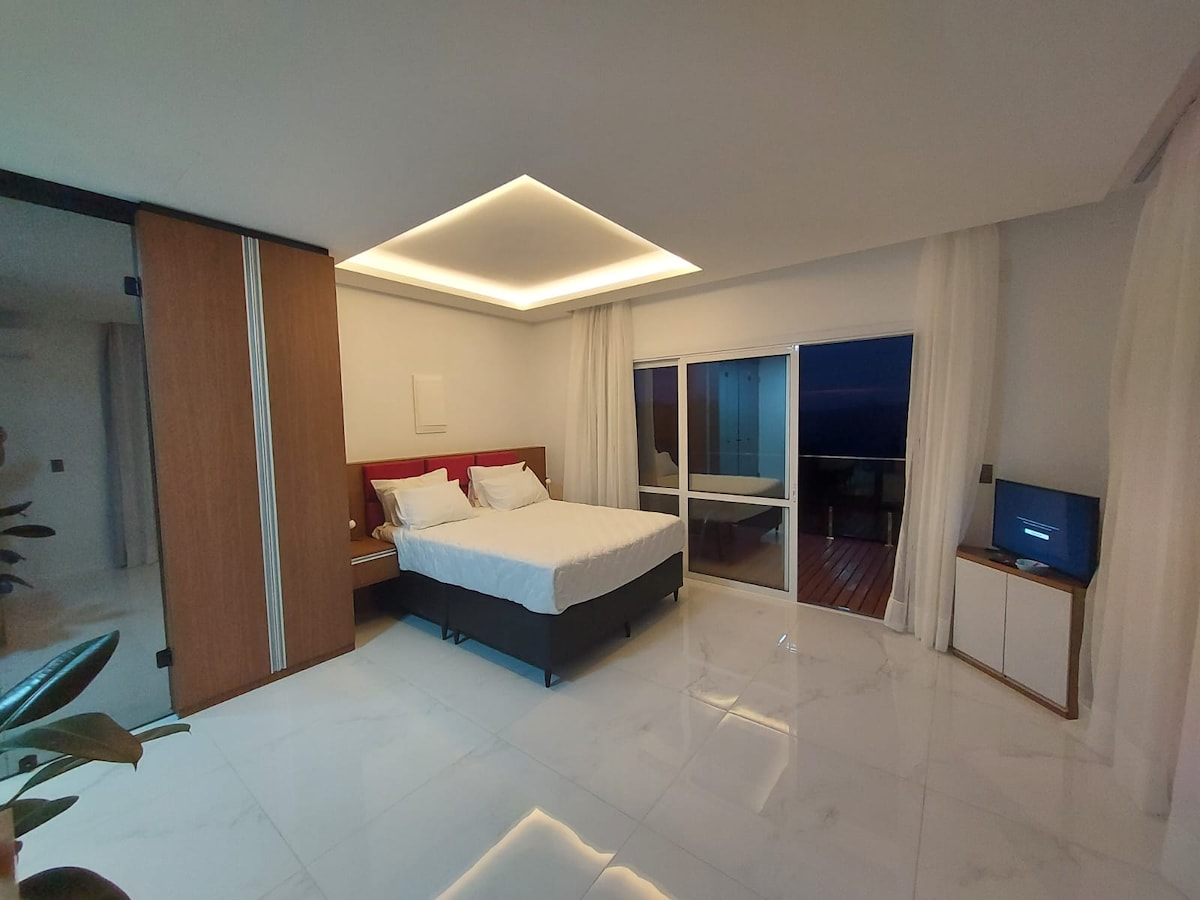A spacious bedroom is equipped with a large bed adorned with white linens. Soft, ambient lighting enhances the room's modern aesthetic, while floor-to-ceiling glass doors offer access to a private deck. Natural light filters through sheer curtains, creating a bright and open atmosphere.