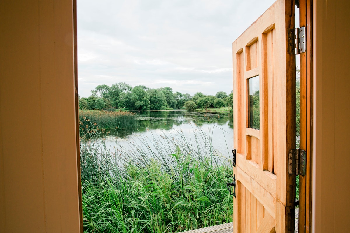The wooden door stands open, revealing a serene view of the lake surrounded by lush greenery. Tall grasses frame the reflective water, creating a peaceful natural setting just beyond the entrance.
