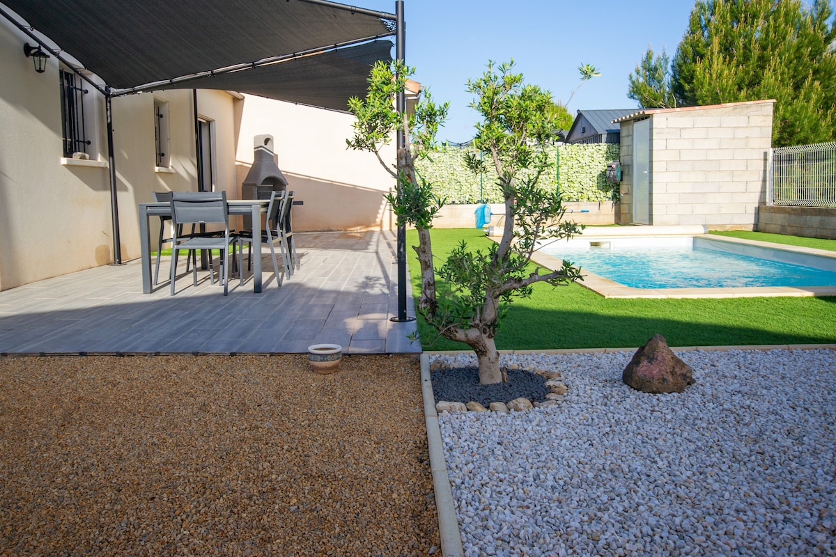A shaded patio area is visible, featuring a dining table with chairs, complemented by a small tree and decorative stones in the garden. The private pool is seen in the background, surrounded by well-maintained grass and landscaping.