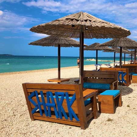 A sandy beach area is shown with several wooden benches featuring blue cushions arranged under straw umbrellas. The calm sea extends in the background, complemented by a clear sky with scattered clouds.